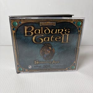 May include: A Baldur's Gate II: Shadows of Amn PC game in a clear plastic case. The game's cover features a teal background with gold lettering and ornate border details. The game title is prominently displayed.
