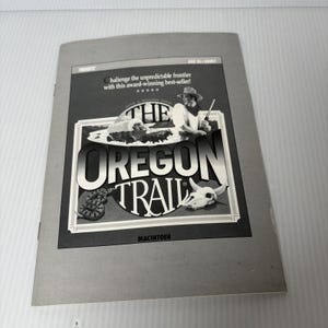 May include: A gray and white vintage Macintosh game box for "The Oregon Trail." The cover features a cowboy illustration, the game's title, and the text "Challenge the unpredictable frontier." The box is slightly worn.