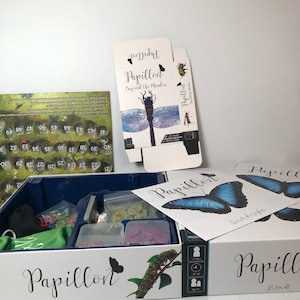 May include: The board game Papillon Beyond the Meadow includes a white box with a butterfly and caterpillar illustration, a game board with numbered spaces, a rulebook featuring a blue butterfly, and plastic containers holding game components.