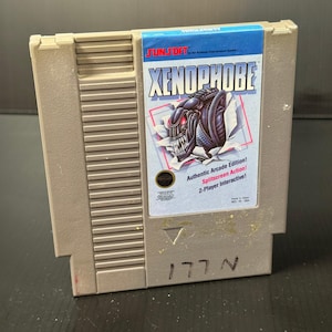 Op de afbeelding: Een beige Nintendo Entertainment System (NES) spelcartridge voor Xenophobe. Het label toont een dreigende alien-illustratie die door gescheurd papier breekt, met de tekst "Authentic Arcade Edition! Splitscreen Action! 2-Player Interactive!"
