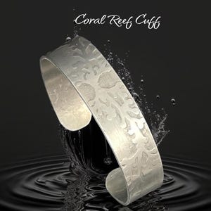 May include: A silver cuff bracelet with a textured coral reef design. The bracelet is open and has a curved shape. The words "Coral Reef Cuff" are written in cursive above the bracelet.