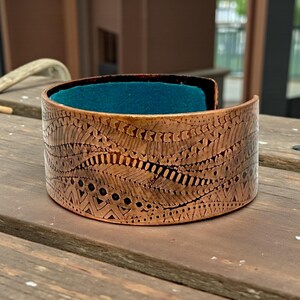 May include: Copper cuff bracelet with intricate etched patterns. The wide band features a detailed design of lines, dots, and geometric shapes. The interior is lined with teal fabric, creating a striking contrast. This handcrafted jewelry piece is a statement accessory.