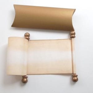 May include: A rolled up scroll with gold-colored end caps and a white background. The scroll is sitting on a gold-colored pillow box.