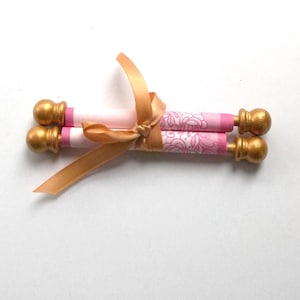 May include: Two rolled paper scrolls tied with a gold ribbon. The scrolls have gold finials and pink and white floral patterned paper. The ribbon is tied in a bow.