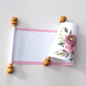 May include: A decorative scroll with a white paper center and pink border. The scroll is held by gold-colored finials. One end of the scroll is rolled, revealing a floral design with pink and burgundy flowers and green leaves.