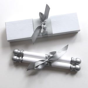 May include: A white rectangular gift box with a silver ribbon tied in a bow, accompanied by two white scrolls featuring silver finials at each end, also tied together with a matching silver ribbon, presented as a set on a plain white background.