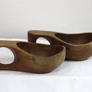 May include: Two dark brown wooden cups with a smooth, rounded bowl shape and a single integrated handle featuring a circular cutout. The wood grain is visible across the surface of both cups, which are positioned side by side on a flat surface.