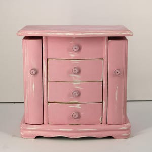 May include: A pink wooden jewelry box featuring four central drawers and two side cabinets. The finish has a distressed appearance with white paint showing through the pink surface. Each drawer and cabinet door has a small, round, matching pink knob.