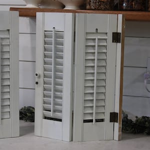 Puede incluir: Un par de contraventanas de madera blanca con lamas horizontales y una varilla de ajuste vertical. Las contraventanas están unidas por bisagras metálicas en el lateral e incluyen un pequeño pomo blanco en el marco, con un acabado pintado.