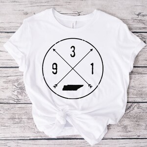 Puede incluir: Camiseta blanca con un diseño circular negro que presenta el estado de Tennessee, flechas cruzadas y los números 3, 9 y 1.
