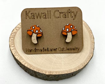 May include: A pair of wooden mushroom stud earrings. The mushrooms are orange with white spots and have a white stem. The earrings are on a brown card with the text "Kawaii Crafty Handmade Laser Cut Jewelry".