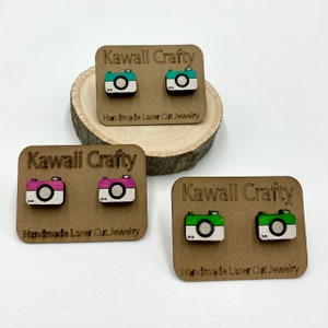 Puede incluir: Tres pares de pendientes de cámara de madera en cartulina marrón con el texto "Kawaii Crafty Handmade Laser Cut Jewelry". Los pendientes son de color verde, rosa y turquesa.