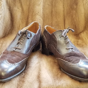 May include: A pair of brown leather and suede lace-up shoes with wingtip detailing. The shoes have a brown leather sole and a brown suede upper.
