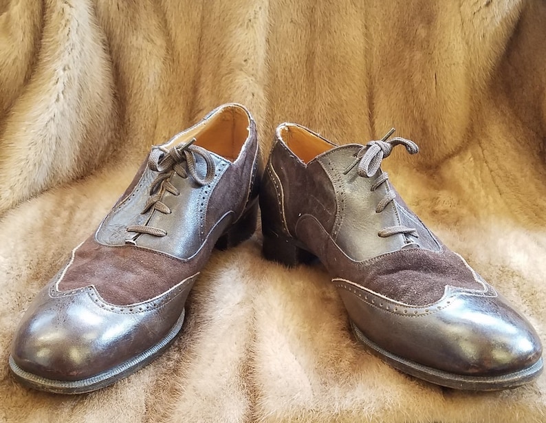 May include: A pair of brown leather and suede lace-up shoes with wingtip detailing. The shoes have a brown leather sole and a brown suede upper.