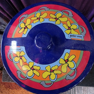May include: A round, blue lid with a central knob and a colorful floral design. The lid features yellow flowers with blue and gray accents, set against a rainbow-colored background. The text "peter max" is visible on the lid.