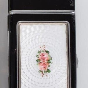 May include: Black rectangular lighter with a silver-colored top and a decorative white panel. The panel features a floral design of pink roses and green leaves. The lighter has a silver-colored metal frame.