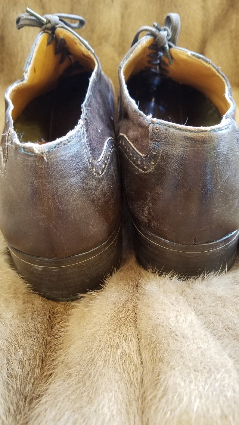 May include: A pair of brown leather shoes with laces. The shoes have a brogue design and are sitting on a light brown fur surface.