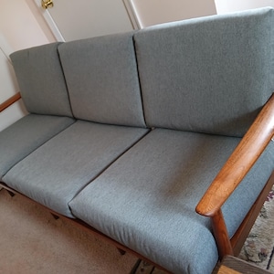 May include: A mid-century modern sofa with a light gray fabric seat and back. The sofa has a wooden frame with armrests. The sofa is a three-seater and is placed on a patterned rug.