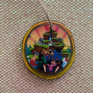 May include: A round wooden ornament with a colorful image of cartoon characters from the movie 'The Fox and the Hound'. The characters are standing on a hilltop with a waterfall in the background. The ornament has a string attached for hanging.