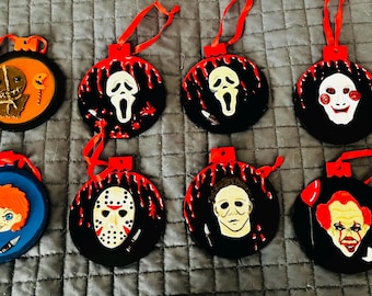 May include: A set of eight circular hanging ornaments featuring various horror movie characters. The designs include Sam, Ghostface, Jigsaw, Chucky, Jason Voorhees, Michael Myers, and Pennywise, all accented with red ribbons for hanging.