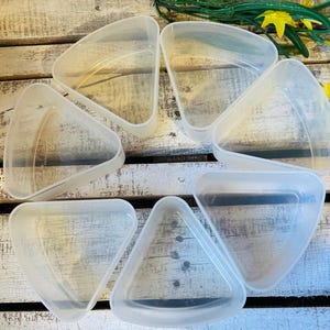 May include: A collection of six clear, triangular plastic containers. The containers are arranged in a circular pattern on a wooden surface. The containers appear to be empty and are translucent. Green and yellow floral accents are visible in the background.