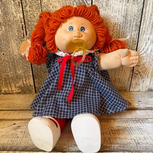 May include: A doll with bright orange yarn hair styled in pigtails, blue eyes, and a tan complexion. The doll wears a navy blue dress with a white grid pattern, a red ribbon tie at the neckline, and a yellow plastic pacifier in its mouth.