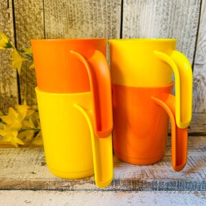 May include: A set of four stackable mugs in bright orange and yellow colors. Each mug features a smooth, cylindrical body and a distinct, elongated vertical handle that extends from the rim to the base, allowing for easy stacking and storage.