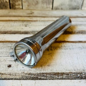May include: A cylindrical metal flashlight with a polished chrome finish. The device features a wide, circular head with a clear glass lens and a reflective interior cone. The body is smooth and tapers slightly toward the base of the handle.