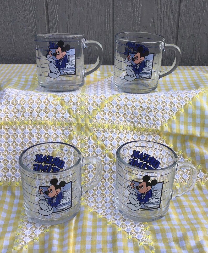 May include: Four clear glass mugs featuring Mickey Mouse in a blue suit, with the words "BREAK TIME" in blue. The mugs have a handle and are decorated with horizontal blue lines.