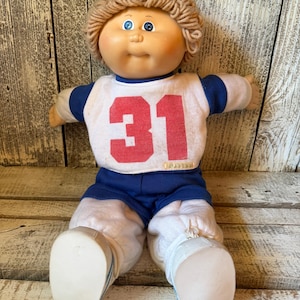 May include: A doll with light brown yarn hair and blue eyes, wearing a white shirt with blue sleeves and the red number 31 printed on the front. The doll is dressed in blue shorts over white pants and white shoes with blue trim on the sides.