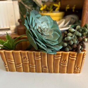 May include: A rectangular, brown ceramic planter with a bamboo design, filled with artificial succulents. The arrangement includes a large, teal-green succulent, small green plants, and a cluster of small, round, green berries.