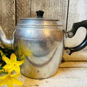May include: A silver-toned metal teapot featuring a rounded body with a decorative beaded band around the upper section, a curved spout, a black lid knob, and a black handle attached with silver rivets, accompanied by bright yellow artificial flowers.