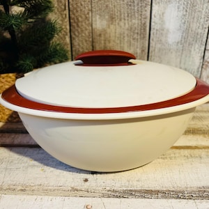 May include: A white serving bowl with a red rim and lid. The bowl has a rounded shape and a handle on the lid. The bowl is sitting on a wooden surface.