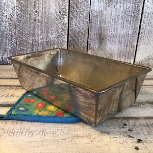 May include: A rectangular, vintage metal loaf pan with a slightly tarnished finish. The pan sits atop a blue and green floral patterned pot holder. The background features weathered, white-painted wooden planks.