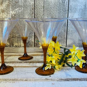 May include: Five clear martini glasses with amber-colored stems and bases are arranged on a white wooden surface. Yellow flowers are placed between the glasses. The glasses have a classic, elegant design, suitable for cocktails.