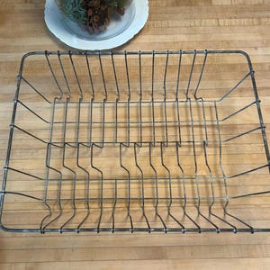 May include: A rectangular, vintage-style wire dish rack with a grid pattern. The rack is made of metal with a weathered, gray finish. The design features vertical and horizontal wires, creating compartments for plates and utensils. The rack sits on a wooden surface.