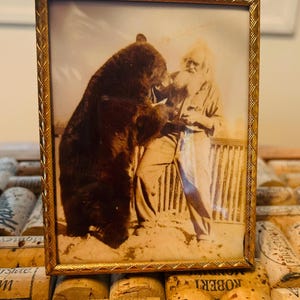 May include: A sepia-toned framed photograph depicts a man interacting with a large bear. The man, with a long white beard, stands facing the bear. The gold-colored frame has a decorative edge. The photograph is resting on a bed of wine corks.