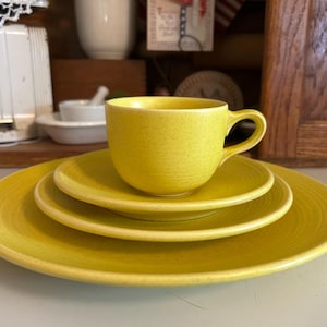 May include: A set of yellow ceramic dishes, including a cup and three plates of varying sizes. The cup sits atop a small plate, with two larger plates beneath. The dishes have a speckled texture.
