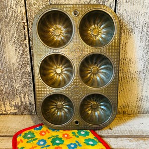 May include: A vintage metal bundt pan with six individual molds. The pan has a textured surface and is marked with the brand name "OVENEX". A yellow and floral patterned pot holder with a red border sits below the pan.