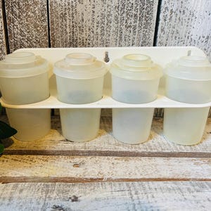 May include: A vintage spice rack with eight translucent plastic containers. The containers are arranged in two rows of four and have matching lids. The rack is white and sits on a weathered wooden surface.