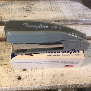 May include: A vintage Swingline 77P stapler in a light blue-grey color, resting on a box of 5000 Standard Staples. The box is white with blue text and a small American flag. The stapler is made in the USA.