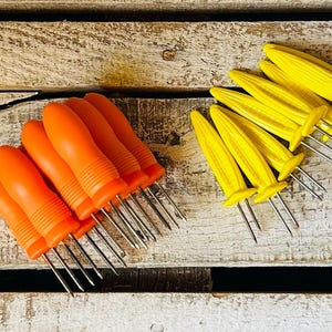 May include: A collection of corn holders in various colors, including yellow, orange, and green. The holders are designed to resemble miniature corn cobs and have metal prongs for easy handling. They are arranged on a weathered, white wooden surface.