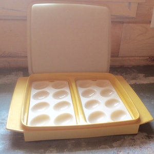 May include: A yellow plastic egg container with a lid. The container has two trays, each holding six egg-shaped indentations. The lid is off, revealing the egg holders. The container is on a gray surface.