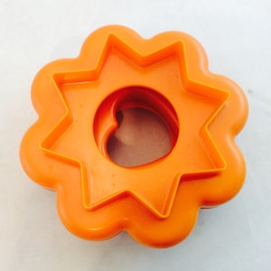 May include: An orange plastic cookie cutter featuring a dual-shape design. The outer edge is shaped like a scalloped flower with rounded petals, while the inner cutting edge forms a sharp, eight-pointed star surrounding a circular center opening.