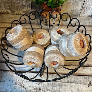 May include: Six balls of beige and white crochet thread are arranged inside a black wire basket. Each ball features a circular red and white label in the center that reads Clark's Big Ball. The thread has a metallic sheen and a tightly wound texture.
