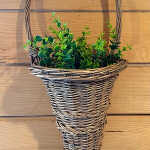 May include: A cone-shaped woven wicker wall basket with a tall arched handle. The basket features a natural light brown finish and is filled with artificial green foliage consisting of small, rounded leaves on thin, branching stems.