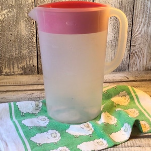 Pu&ograve; includere: Una caraffa in plastica traslucida con coperchio rosa e manico laterale. La caraffa &egrave; dotata di un beccuccio per versare ed &egrave; posizionata su un panno in tessuto bianco e verde decorato con un motivo ripetuto di illustrazioni di pecore bianche.