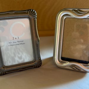 May include: Two small rectangular picture frames. The left frame is dark metallic with shell-like corner details and contains a paper insert reading 2 x 3 For Your Vertical or Horizontal Photo. The right frame is silver with a smooth, rounded border.