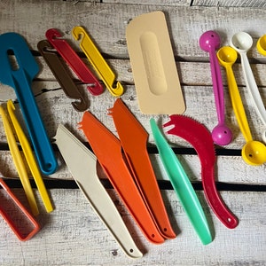 May include: A collection of colorful vintage kitchen utensils, including spatulas, knives, and measuring spoons. The tools are in various colors such as blue, orange, yellow, and red. The items are arranged on a white wooden surface.