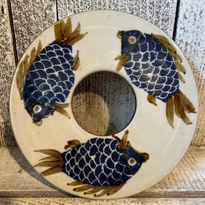 May include: A round, off-white ceramic plate with a central hole, decorated with three blue fish. The fish have detailed scale patterns and brown fins. The plate has a rustic, handmade appearance, suitable for serving or display.
